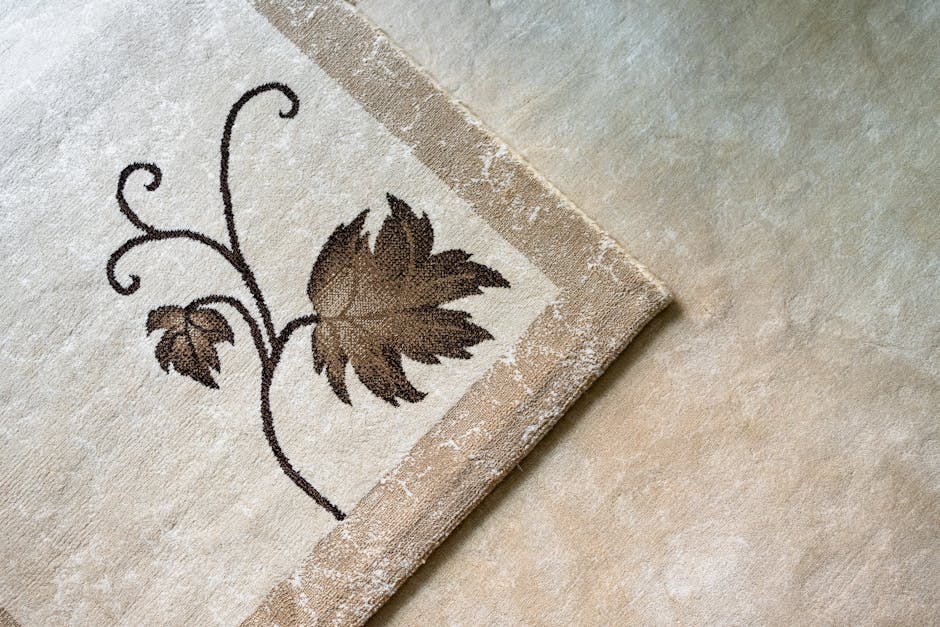 Close-up of a black vacuum cleaner with a hose attachment being used to clean an ornate, patterned area rug on a wooden floor in a residential setting. The rug features a floral design with muted past
